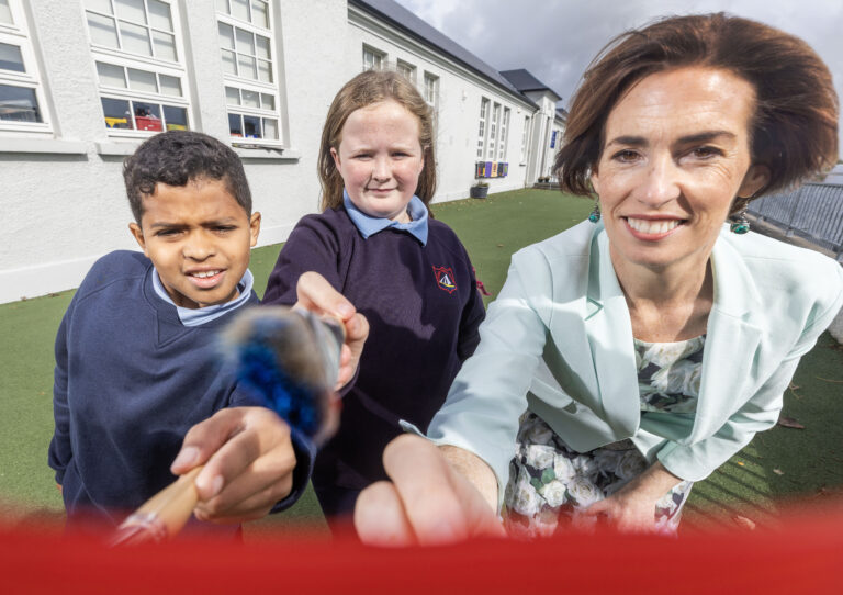 Minister Naughton and pupils reach towards the camera lens with paint brushes.